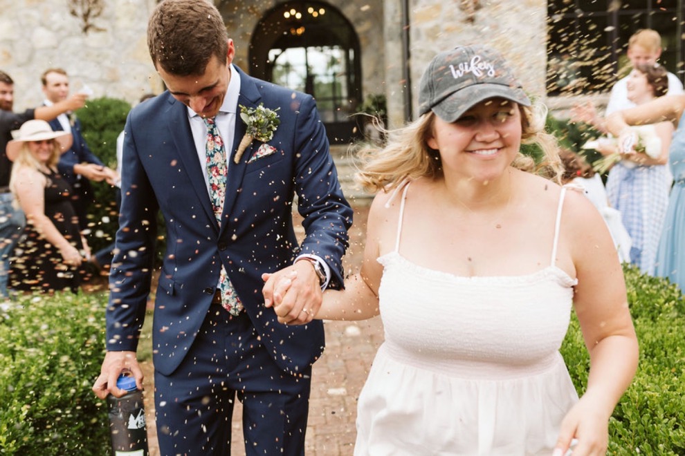 bride and groom running down brick path as guests toss birdseed. Bride wears a white sundress and baseball cap.