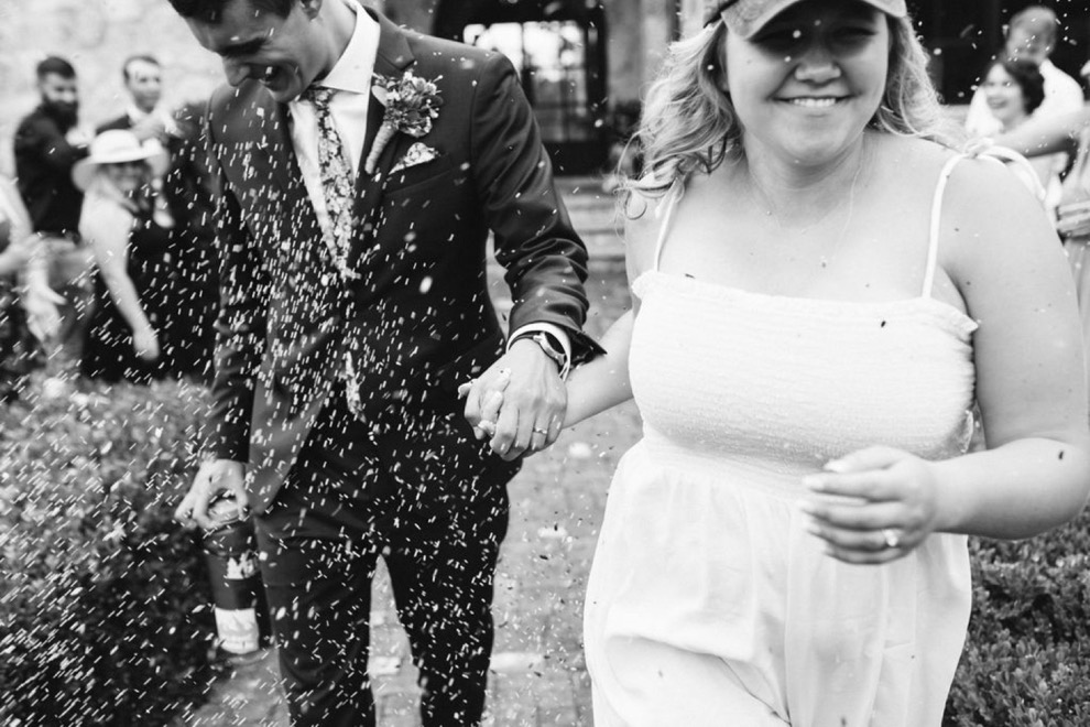 bride and groom running down brick path as guests toss birdseed. Bride wears a white sundress and baseball cap.