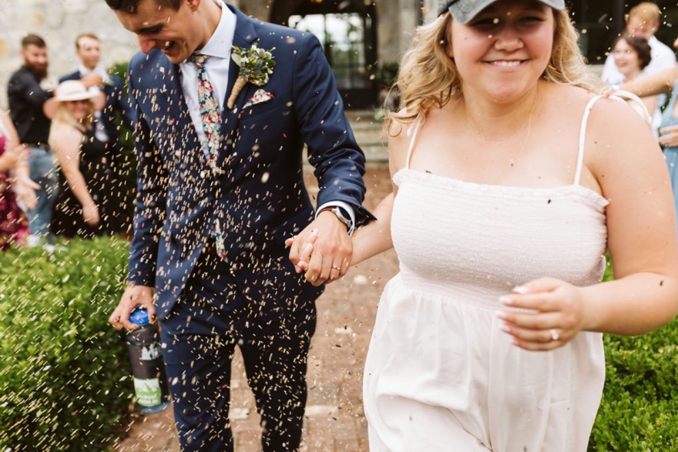 bride and groom running down brick path as guests toss birdseed. Bride wears a white sundress and baseball cap.