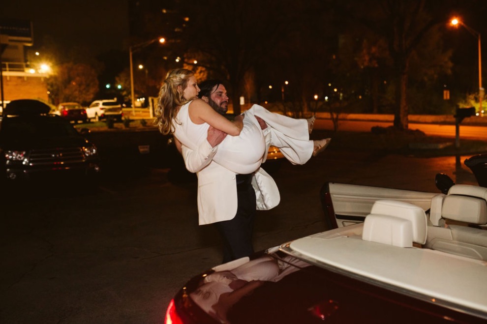 groom carries bride to getaway car after wedding reception
