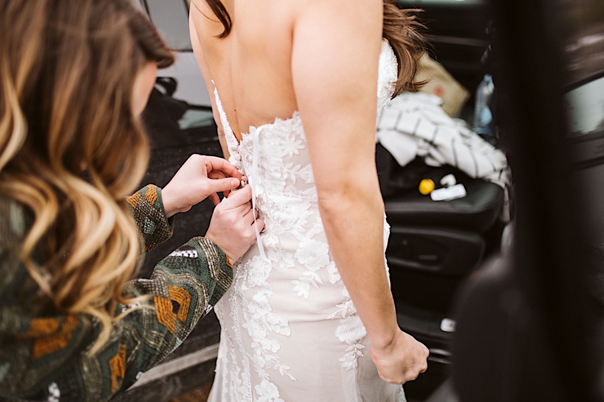 A friend buttons the back of bride's strapless lacy dress next to a car