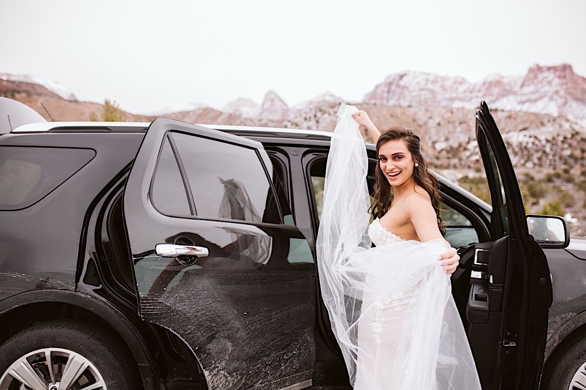 Bride stands in her strapless lacy gown beside black car holding her veil high overhead