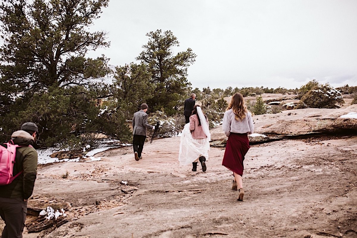 Bride and a few friends hike up a large rock toward scraggly pine trees and shrubs