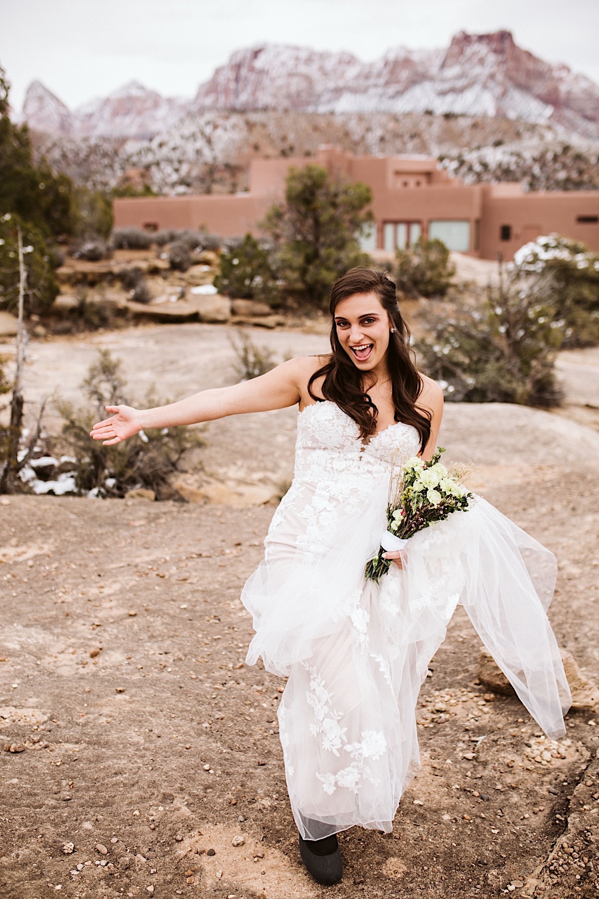 Bride in strapless, lacy dress holds her bouquet with other arm open wide. She lifts her gown to reveal snow boots