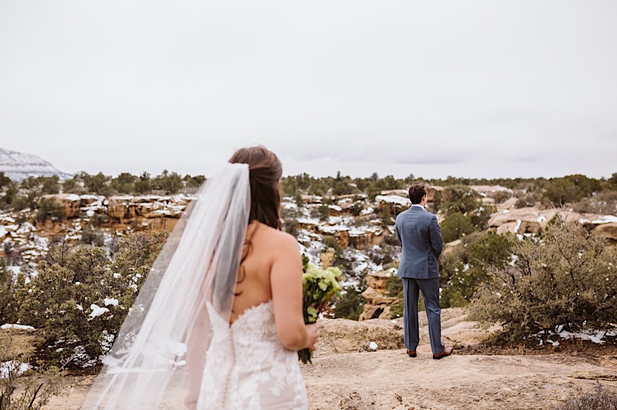Groom stands overlooking a cliff. Bride wearing lacy strapless dress and long veil walks toward him for private first look