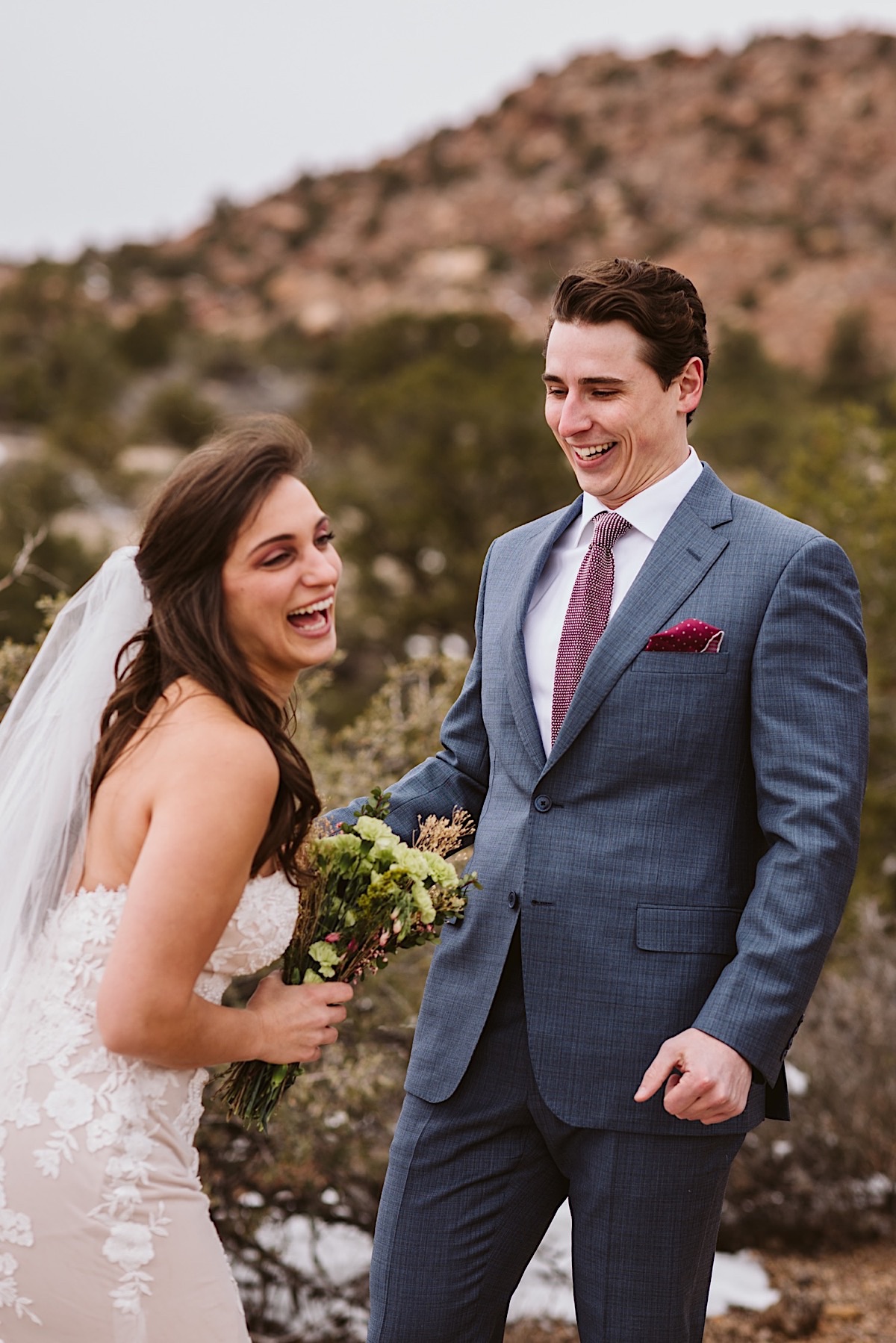 Groom wearing a blue suit with red polka dot tie and pocket square laughs with bride