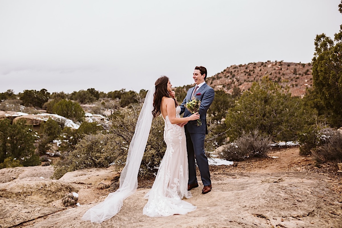 Groom wearing a blue suit with red polka dot tie and pocket square holds bride wearing a strapless lace gown and long veil