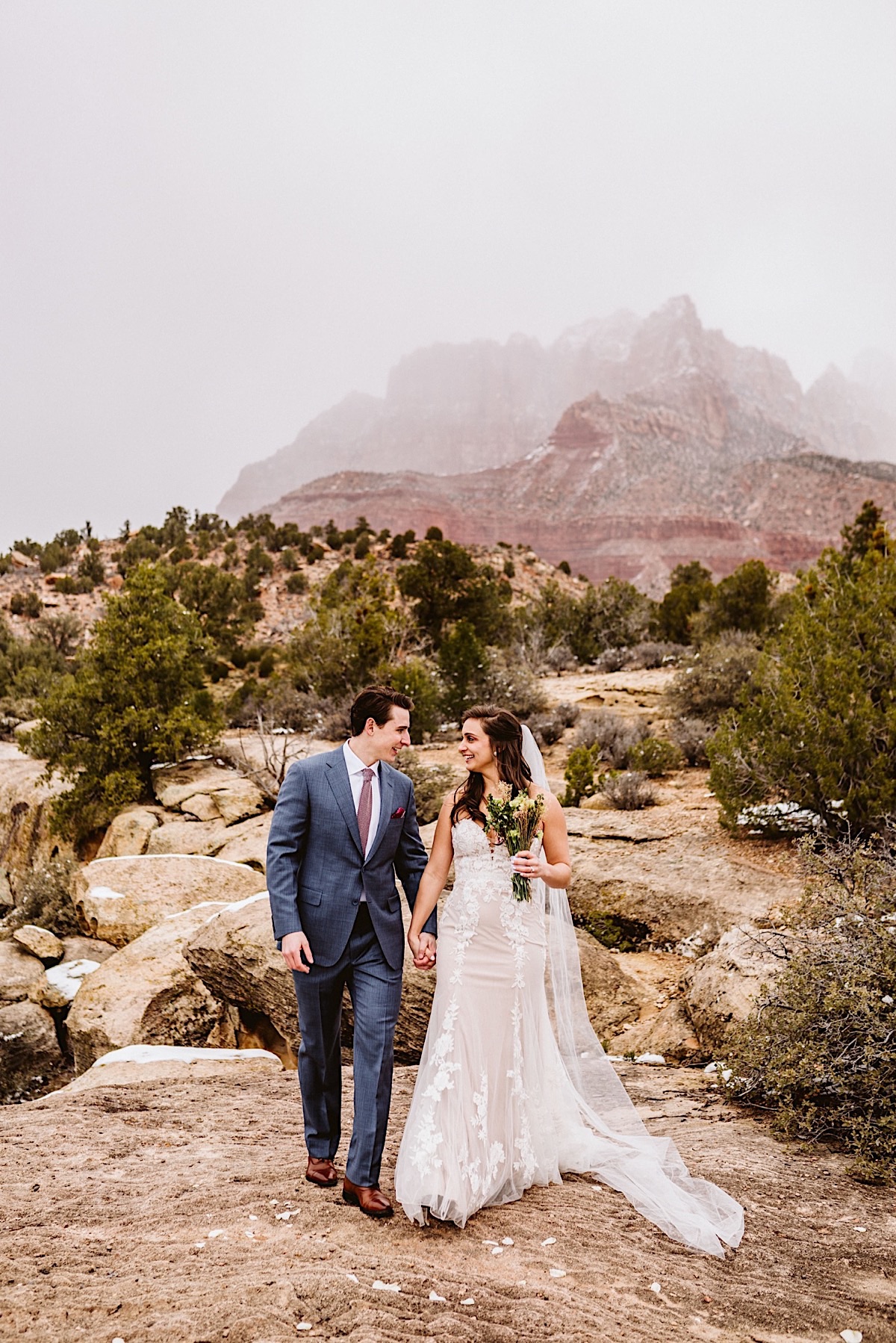 Bride and groom holding hands on the rocks of Zion National Park, snowy fog rolling over the red cliffs in the background
