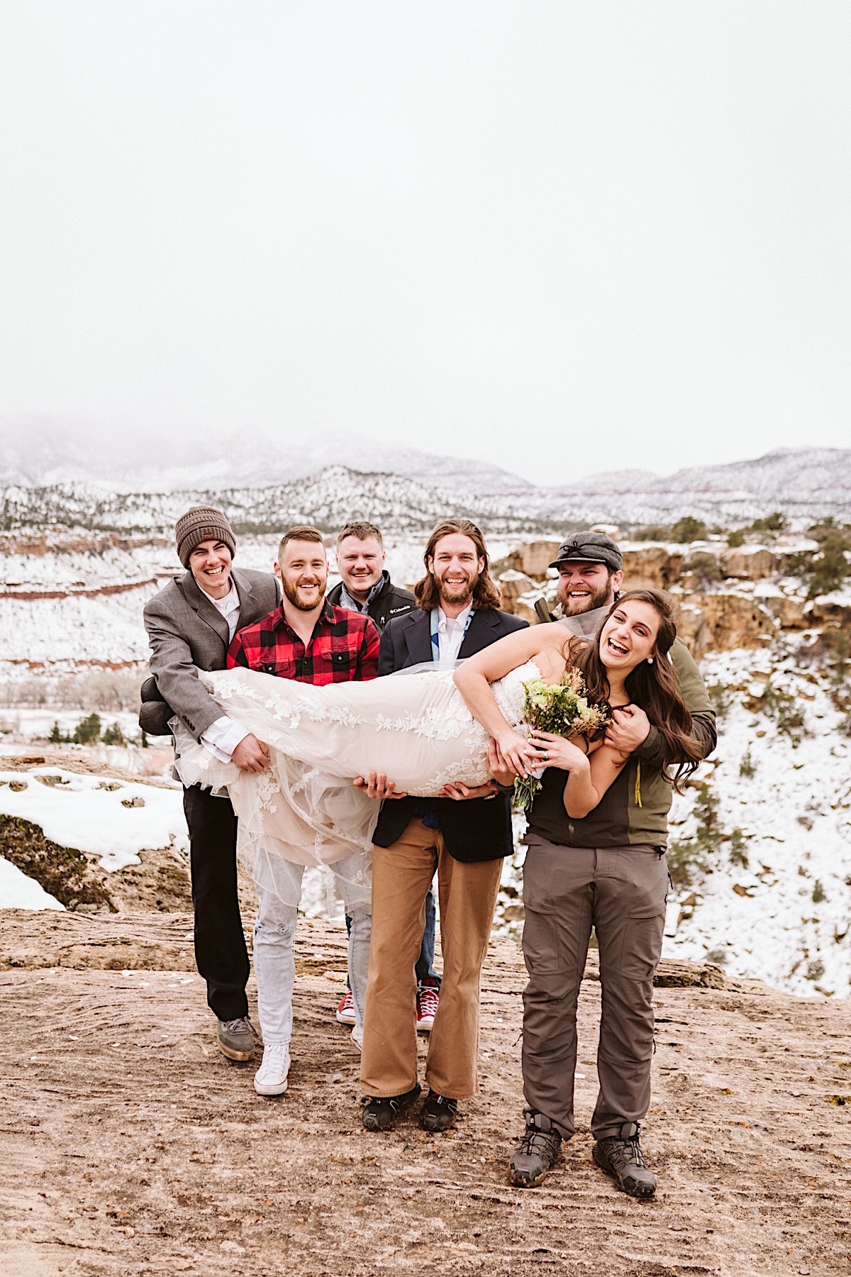 Groomsmen hold the bride across their arms, the snowy hills and cliffs of Zion National Park in the background.