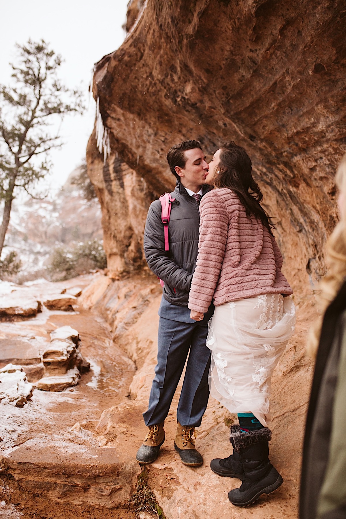 Bride and groom, wearing hiking boots, stop for a kiss next to rock cliffs.