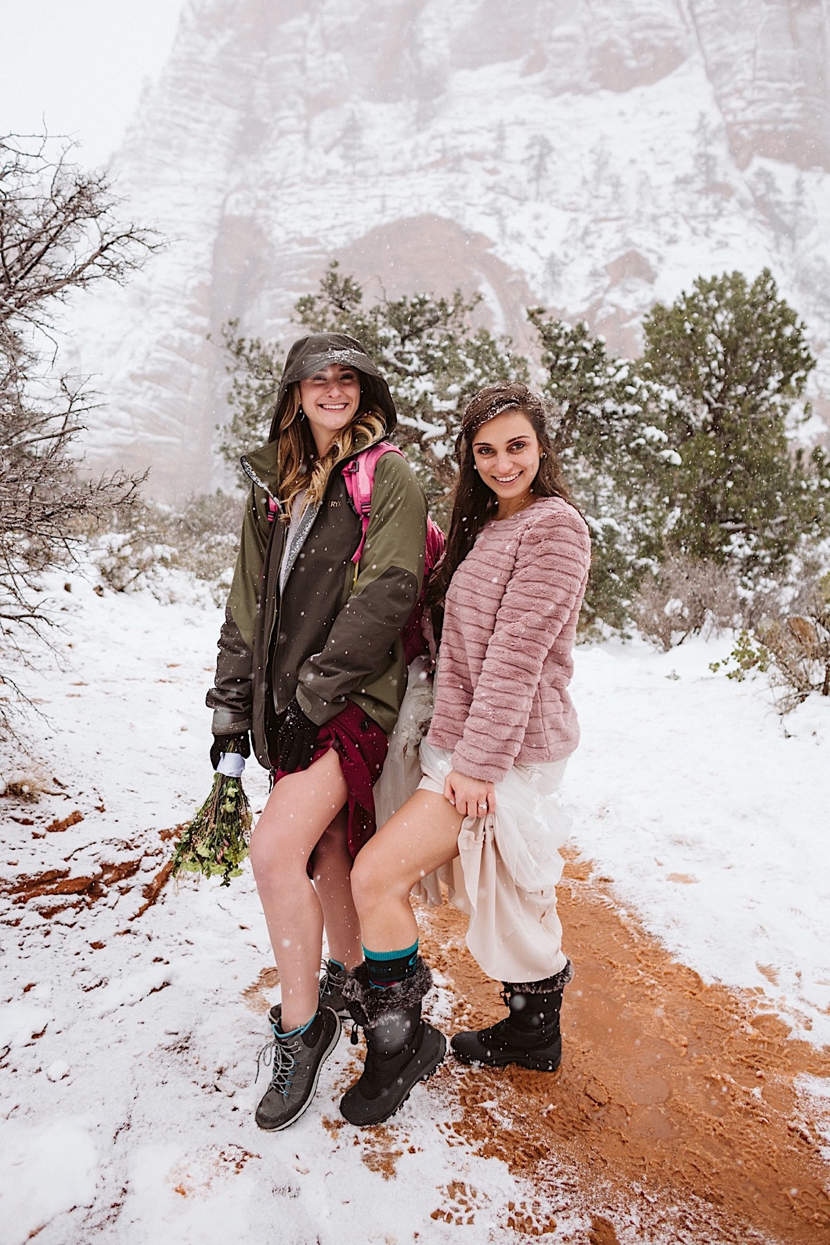 Bride and bridesmaid pull up their dress hems during snowy Zion hike to reveal their hiking boots.