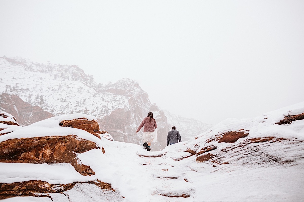 Bride and groom hiking a snowy trail to Canyon Overlook as snow falls around them.