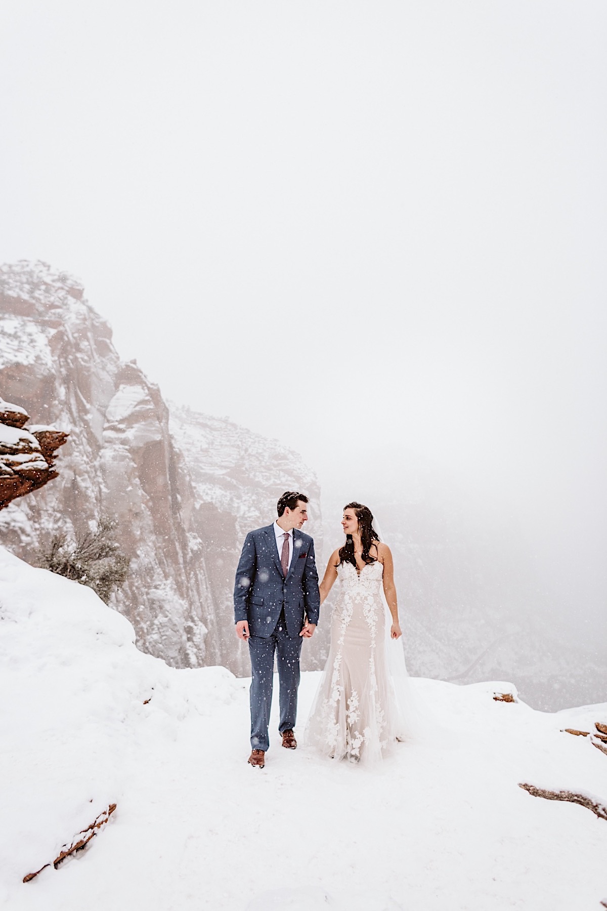 Bride and groom hold hands and look at each other at Canyon Overlook as snow falls around them.