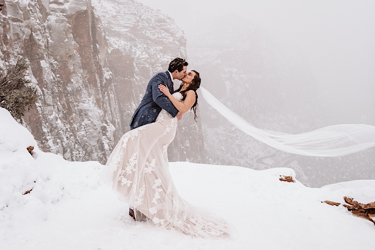 Groom dips bride for a kiss in front of red cliffs as snow falls around them. Brides veil streams behind her.