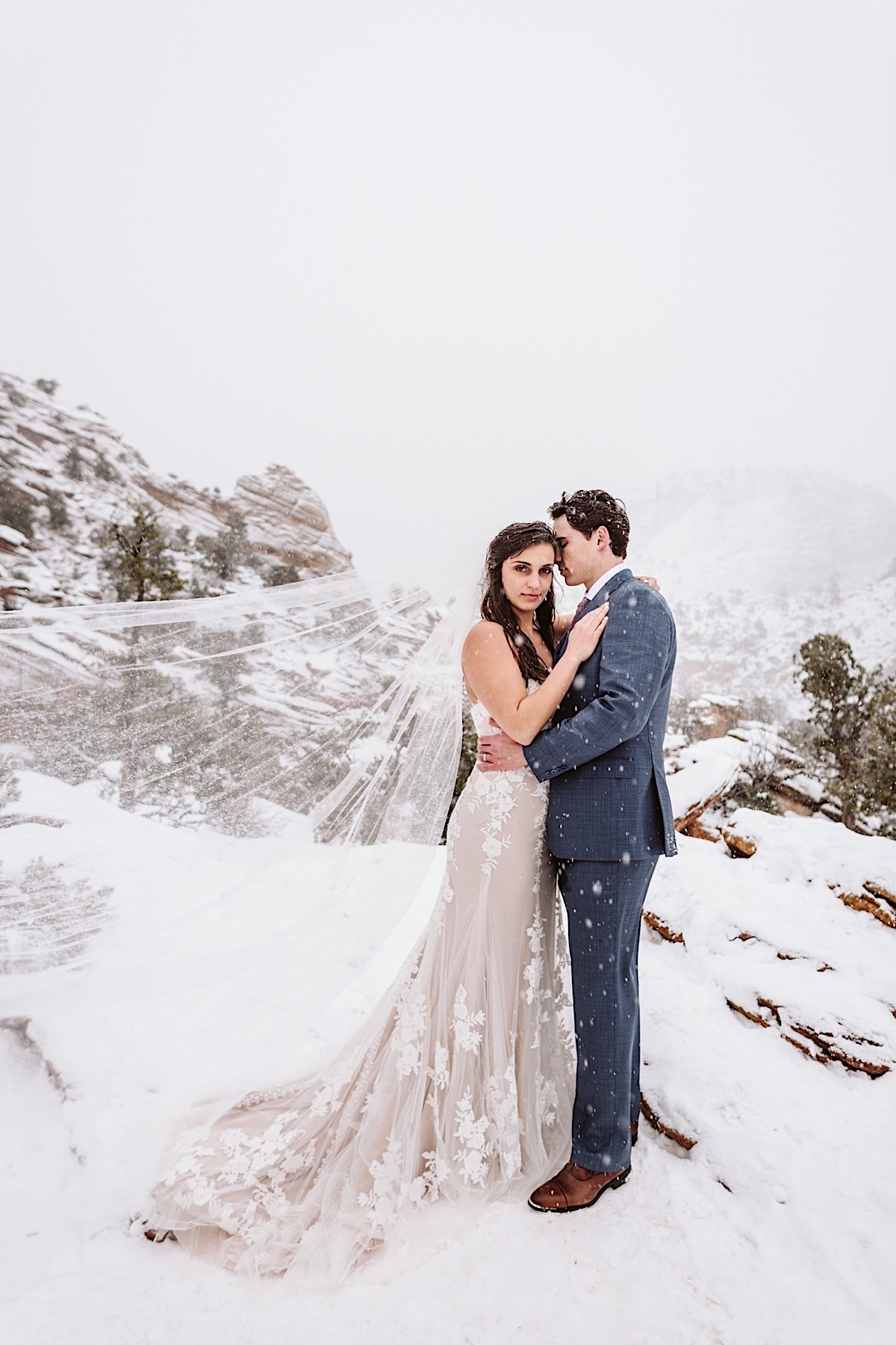 Bride and groom hold each other in front of red cliffs as snow falls around them. Brides veil streams behind her.