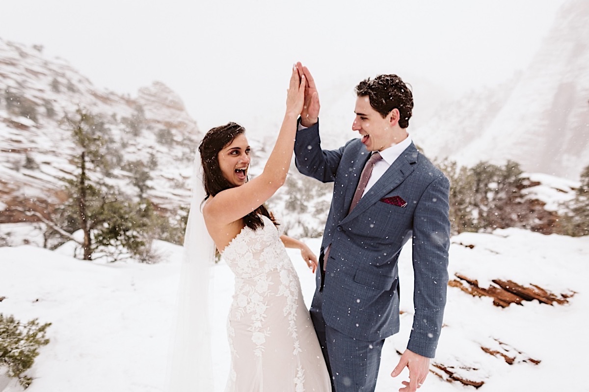 Bride and groom high-five each other, laughing as snow falls around them.