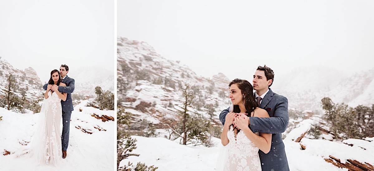 Groom stands behind bride, his arms wrapped around her shoulders and her hands holding onto his hands. Snow falls around them.