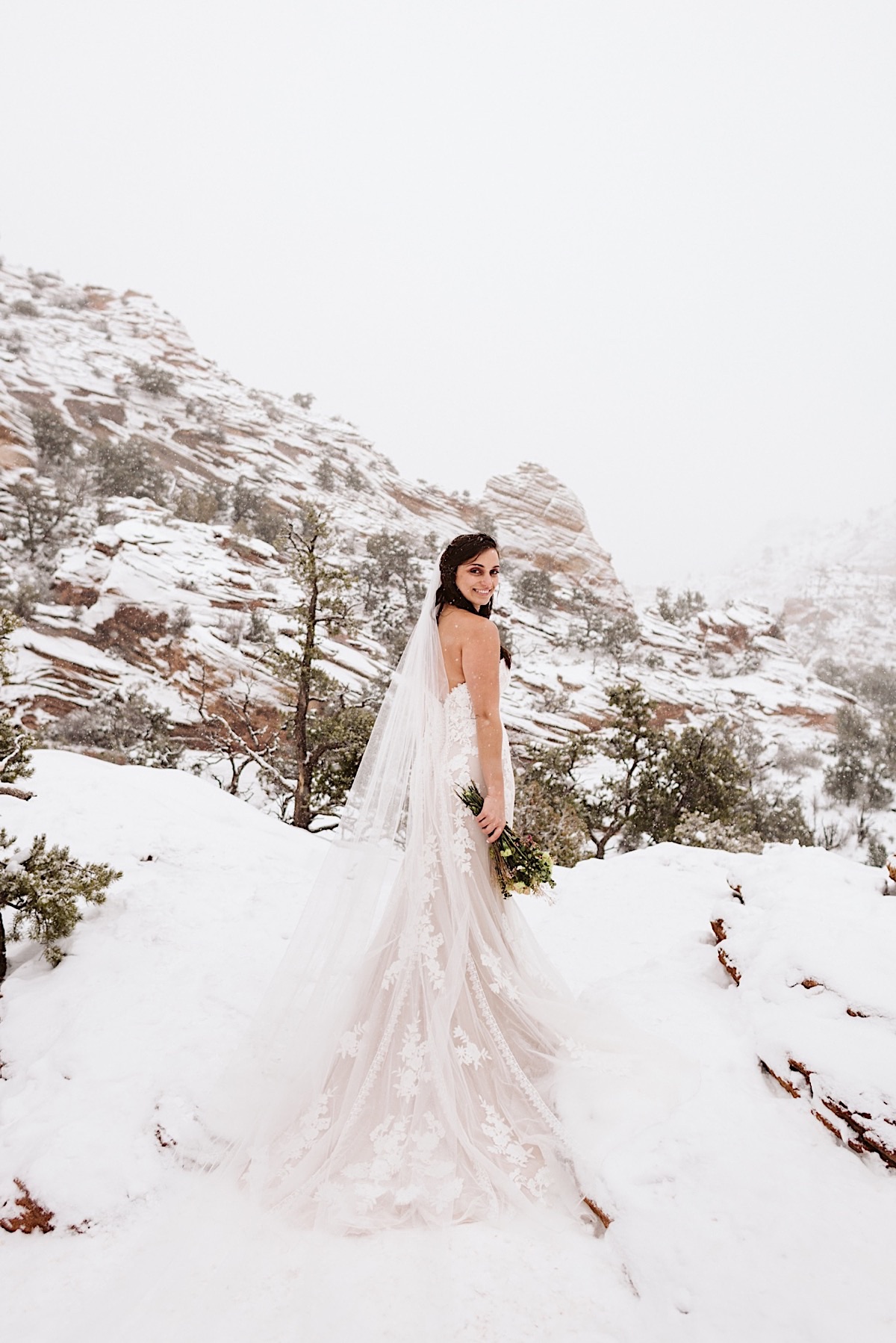 Bride stands on a snowy trail with the red cliffs of Zion behind her. She holds her bouquet at her side.