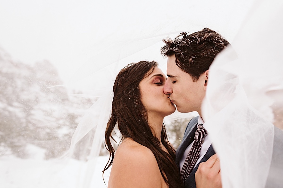 Bride and groom kiss in a snowstorm, her veils swirls around their shoulders.