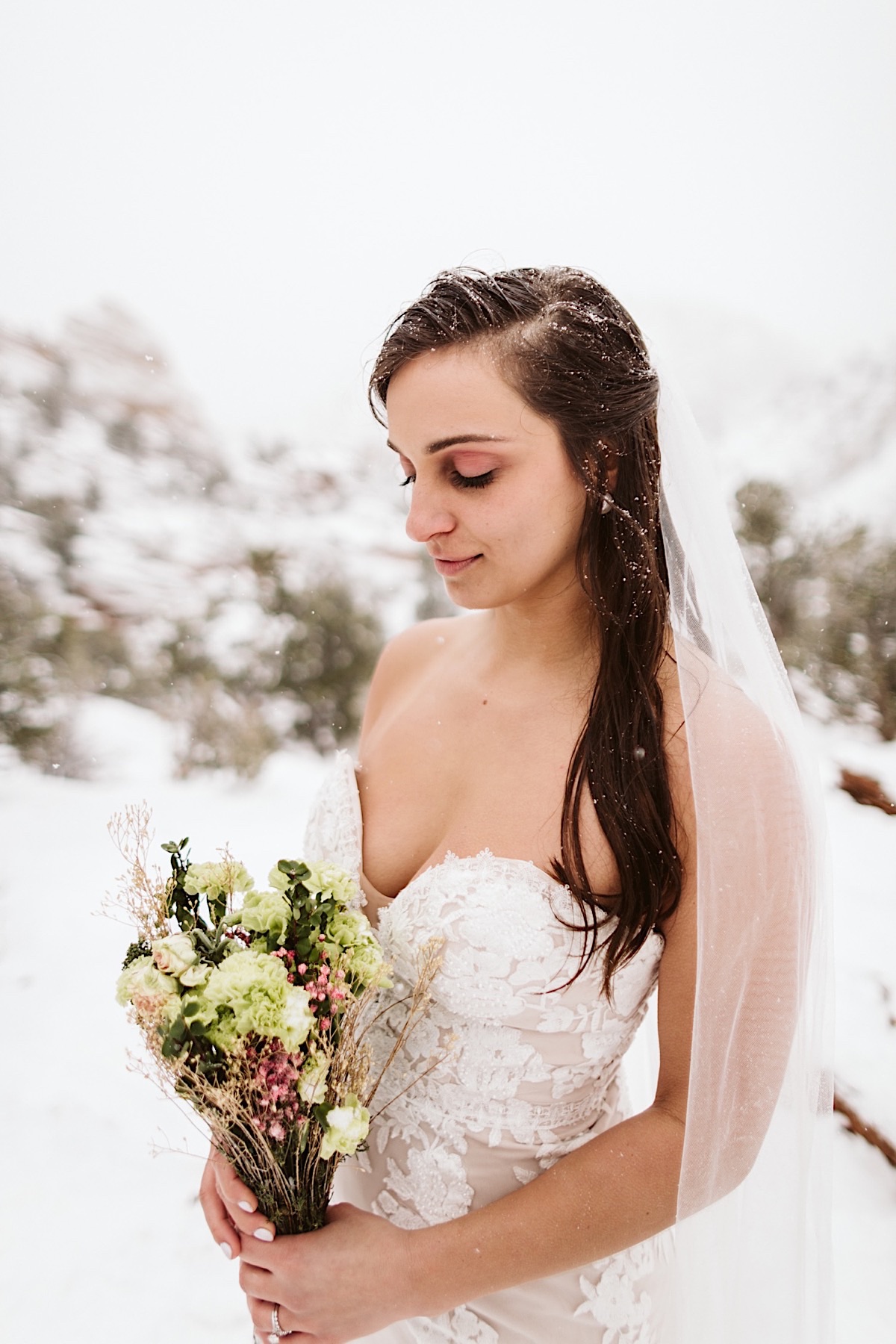 Bride in lace gown looks down at her bouquet of white and purple flowers and greenery. Her dark hair is dusted with snow.