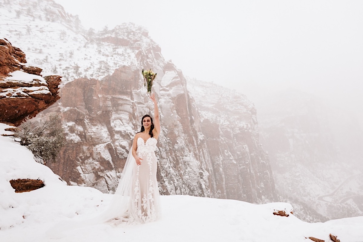 Bride stands in the snow with cliffs barely visible behind her. She tosses her bouquet over her shoulder into the cavern.