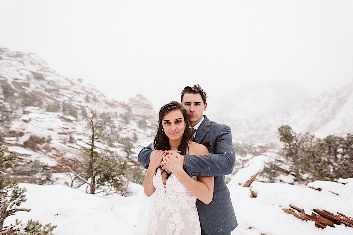 Groom stands behind bride, his arms wrapped around her shoulders and her hands holding onto his hands. Snow falls around them.