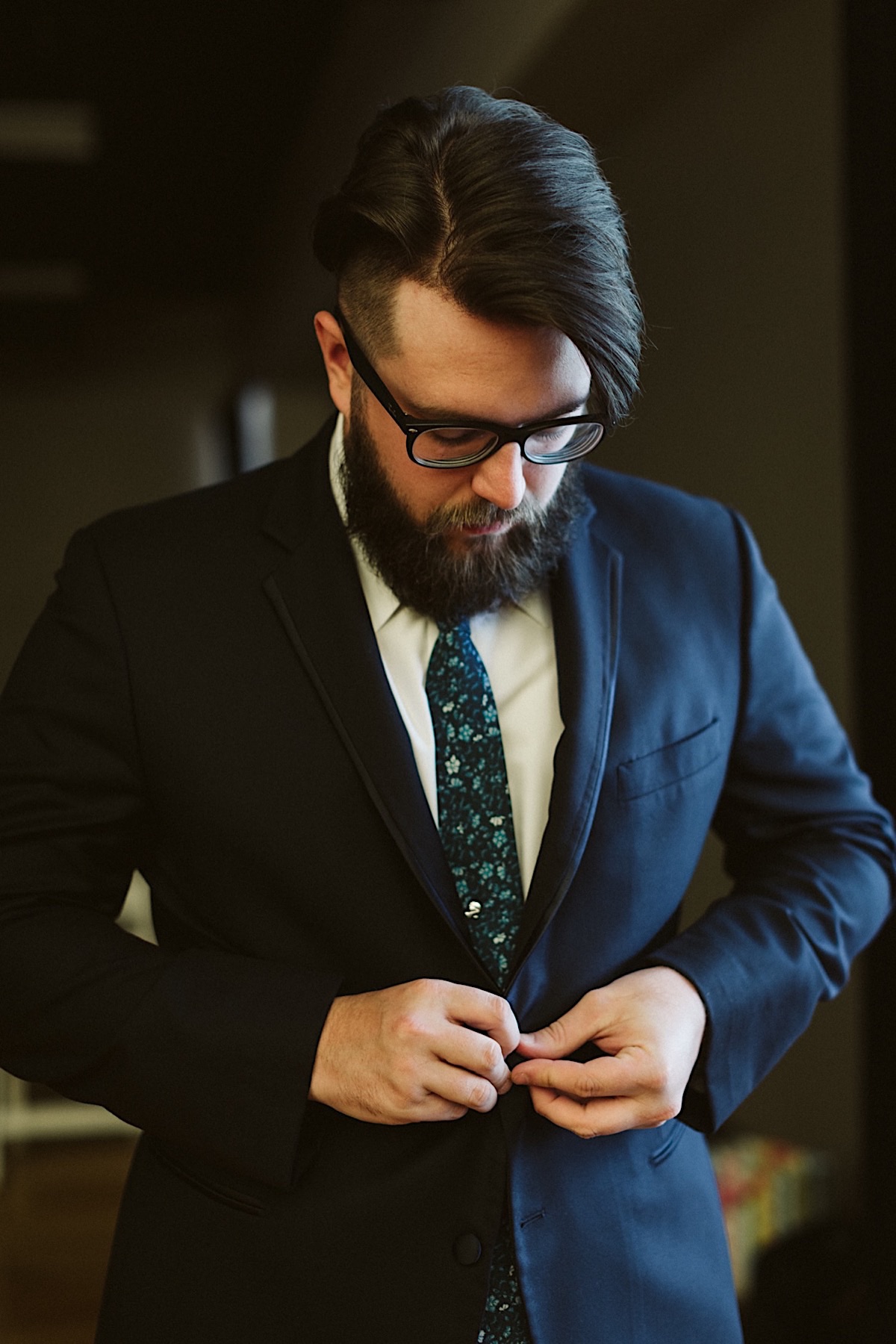 Man wearing glasses buttons his dark suit with blue mosaic tie contrasting against white shirt