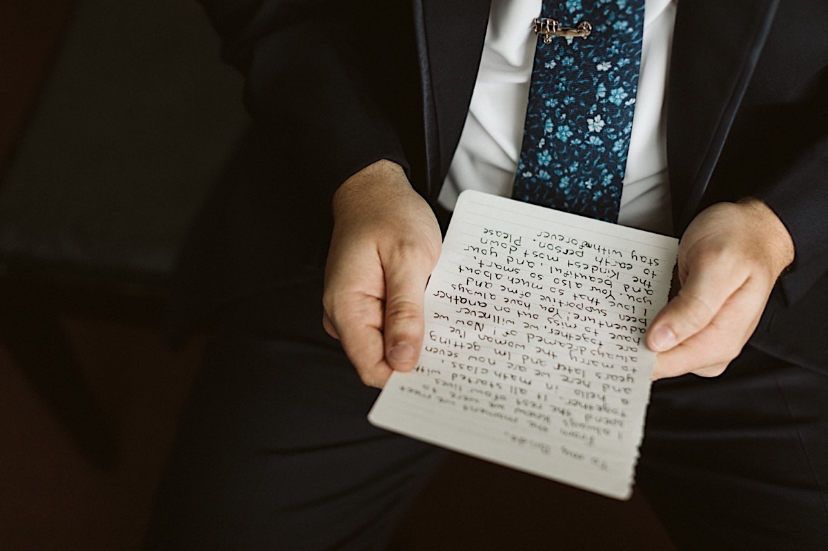 Man holds paper with his wedding vows. His dark suit and blue floral mosaic tie contrast.