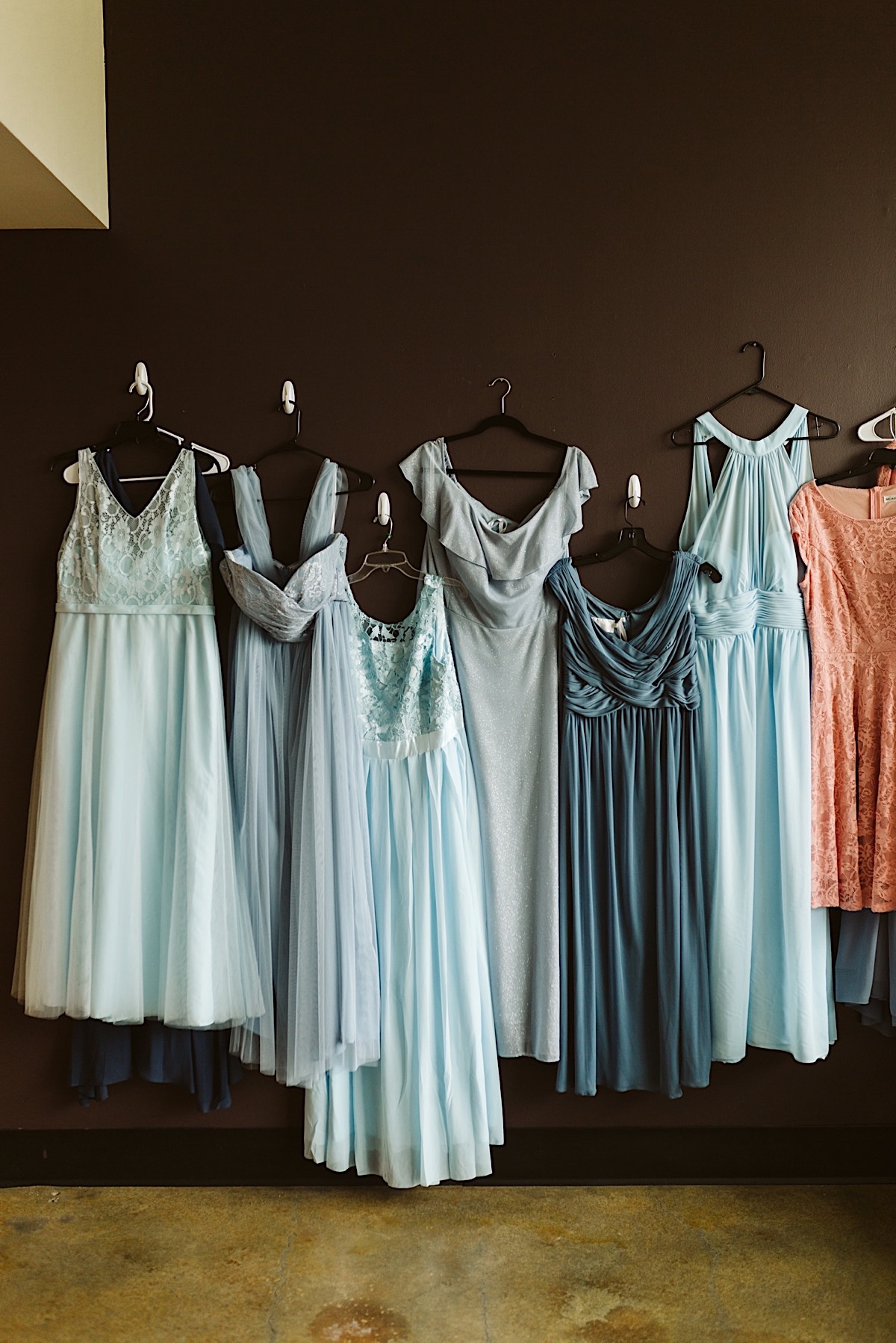 Bridesmaids dresses in various blue tones hang on hooks on a deep brown wall