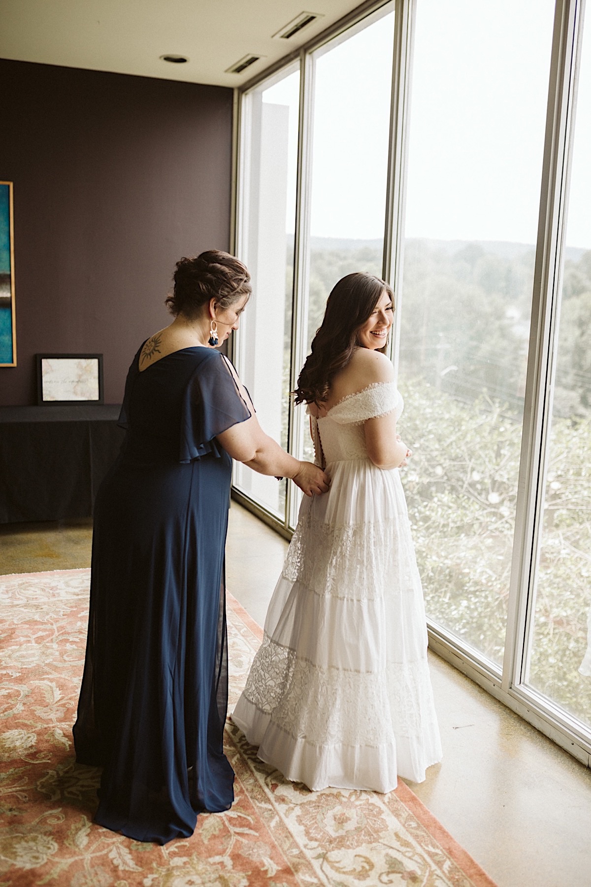 Mother of the bride stands behind her daughter, zipping her lace off-the-shoulder gown. Floor-to-ceiling windows frame them.