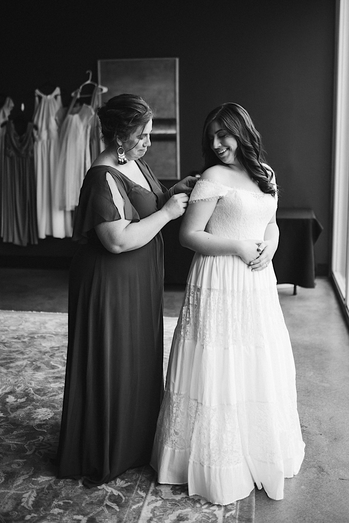 Mother of the bride stands behind her daughter, straightening her lace off-the-shoulder gown