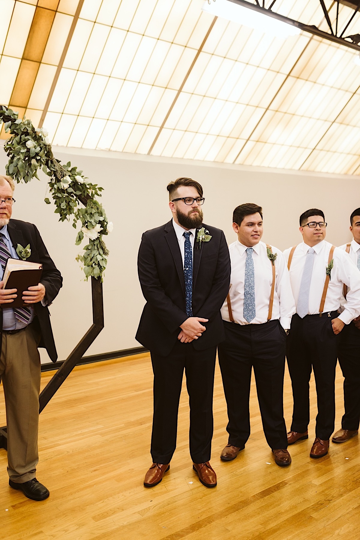 Groom and groomsmen stand under a large skylight near an octagonal wedding arch