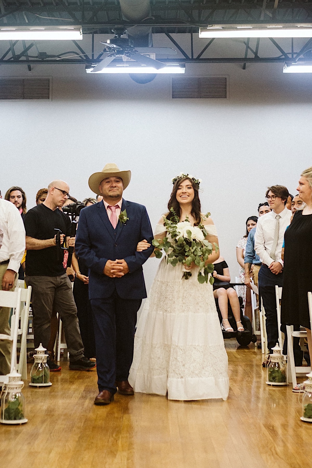 Bride walks down the aisle, escorted by her father who wears a cowboy hat.