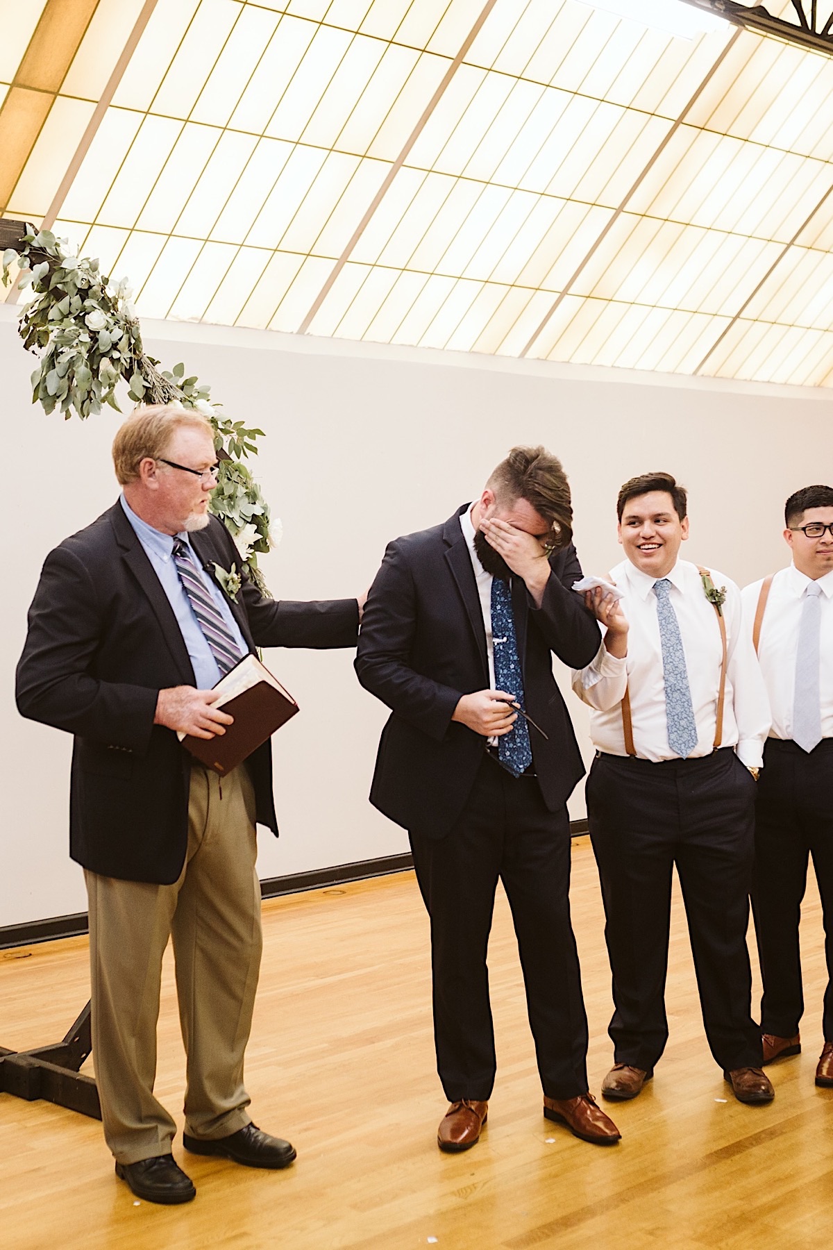 Officiant and best man reach toward groom who is wiping tears from his eyes.