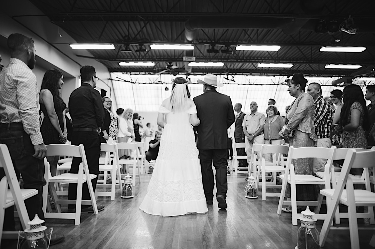 Bride walks down the aisle, escorted by her father who wears a cowboy hat.