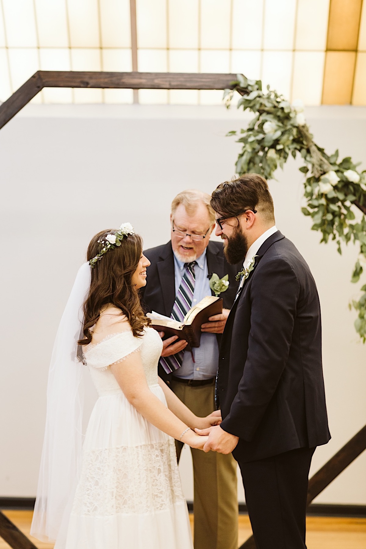 Bride and groom face each other, holding hands under octagonal wedding arch, the officiant reading behind them.