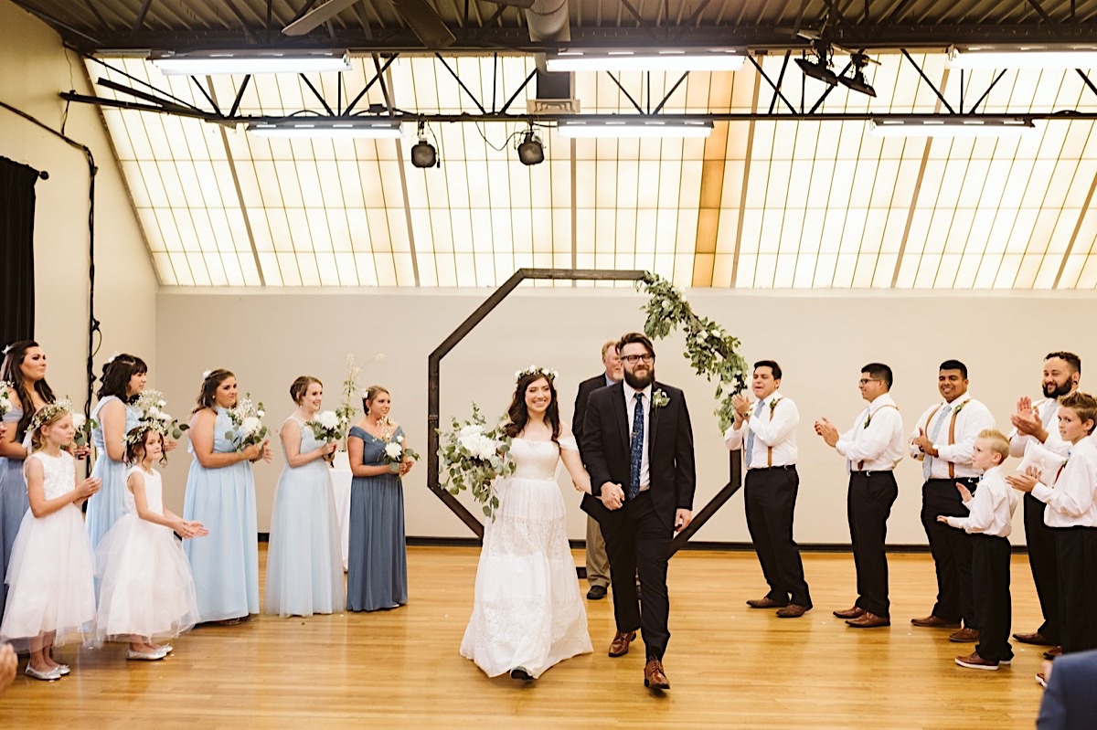 Bride and groom under octagonal wedding arch face their guests. Bridesmaids and groomsmen clap and cheer behind them