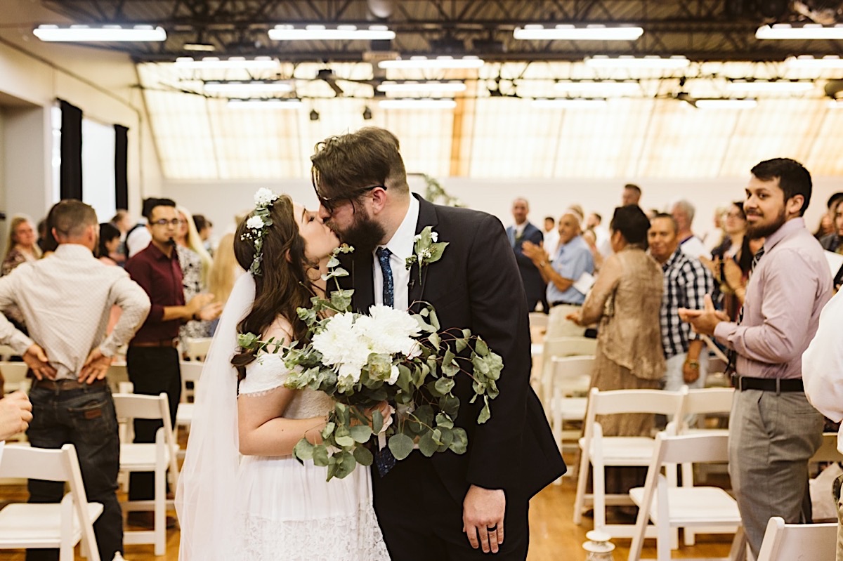 Bride and groom kiss with guests clapping behind them. She holds a white flower and eucalyptus greens bouquet.