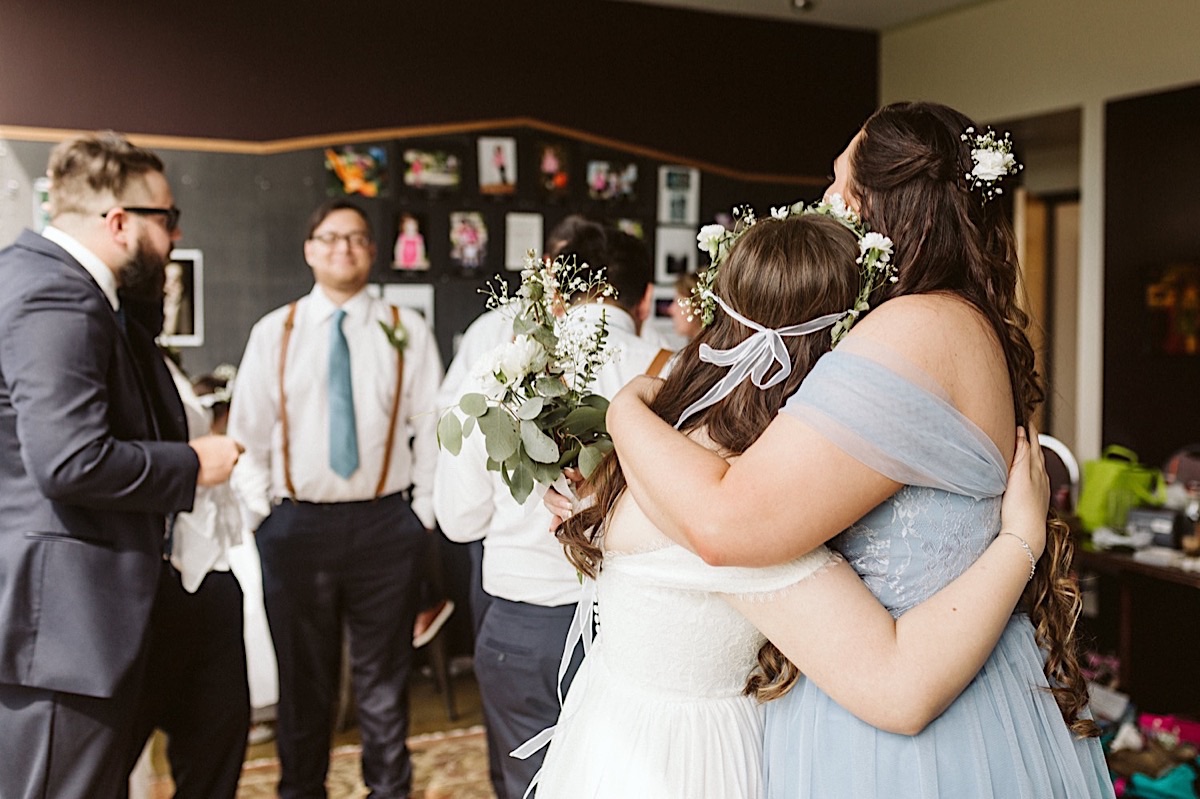 Bride in white gown hugs bridesmaid in blue dress, white flower in each of their dark hair.