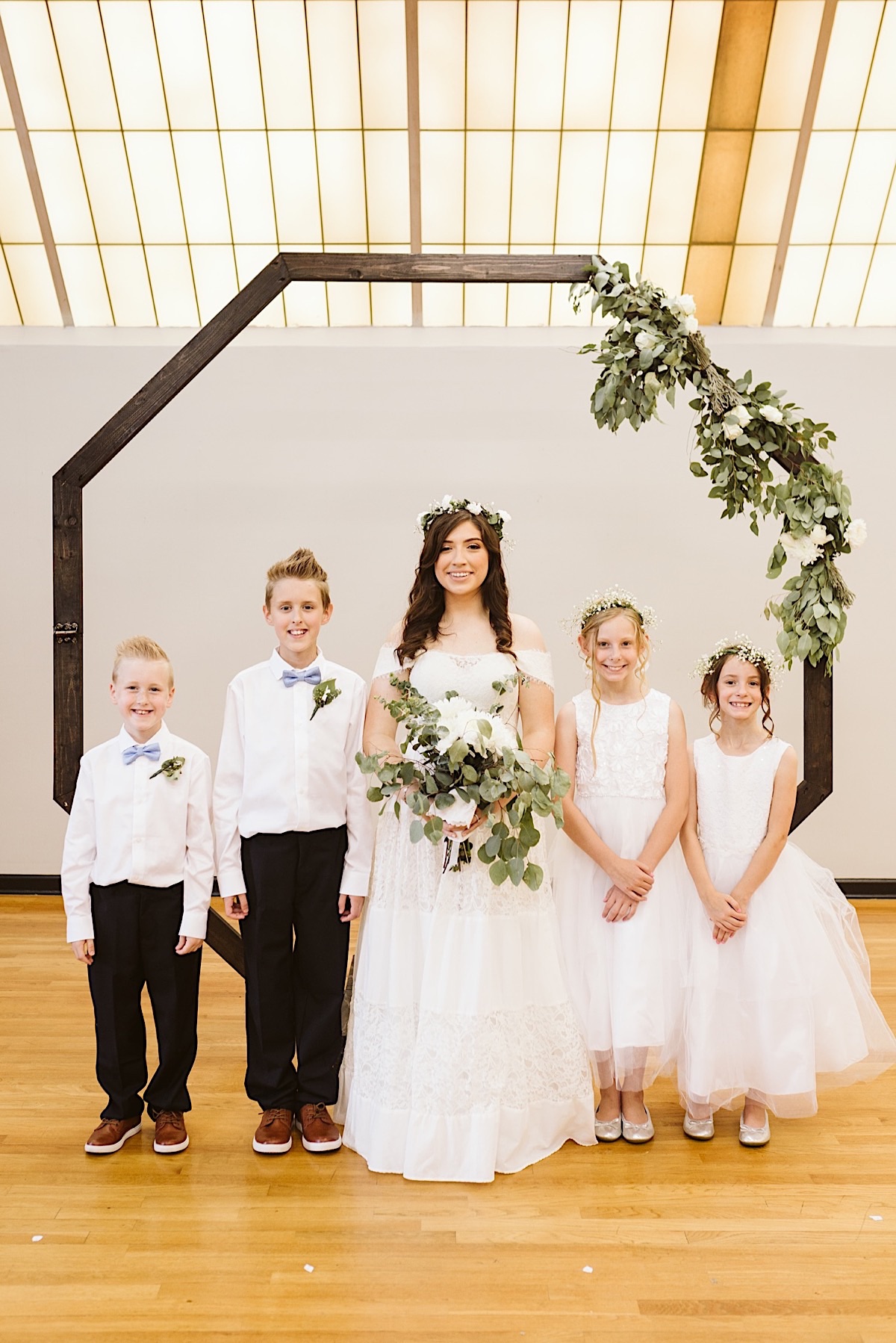 Bride with white and green bouquet stands with ring bearers and flower girls in front of an octagonal wedding arch.