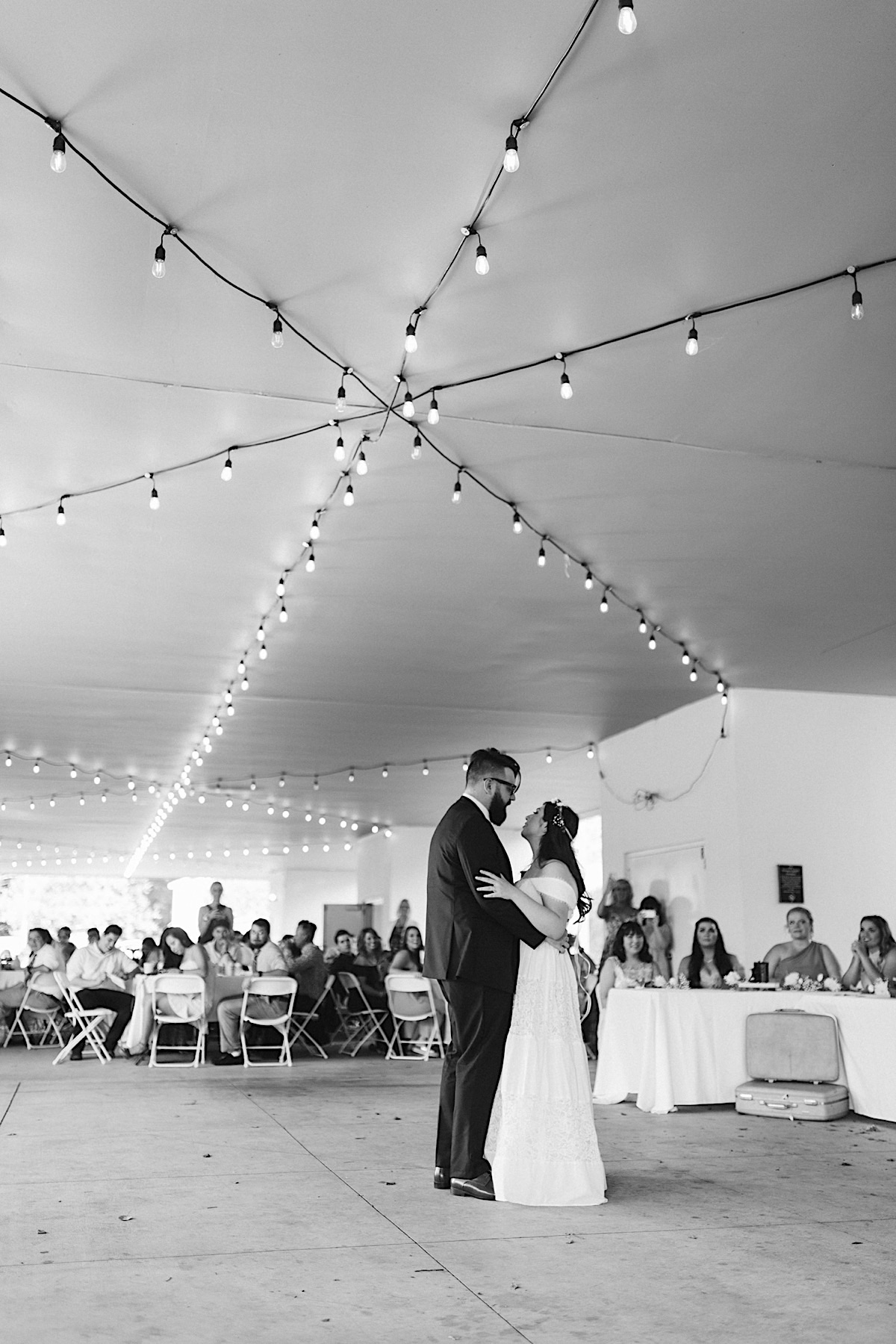 Husband and wife first dance during wedding reception. She cuddles into his chest under white string patio lights