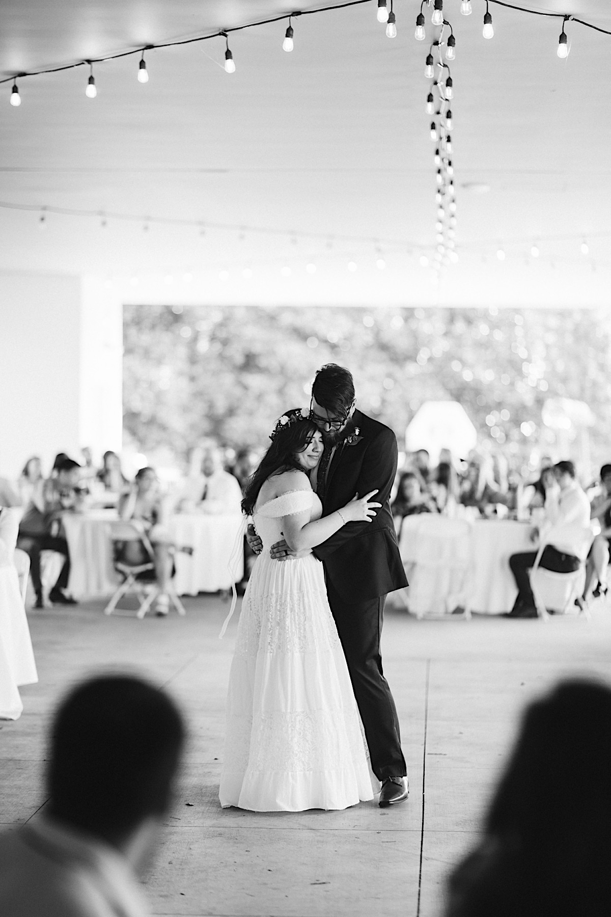 Husband and wife first dance during wedding reception. She cuddles into his chest under white string patio lights
