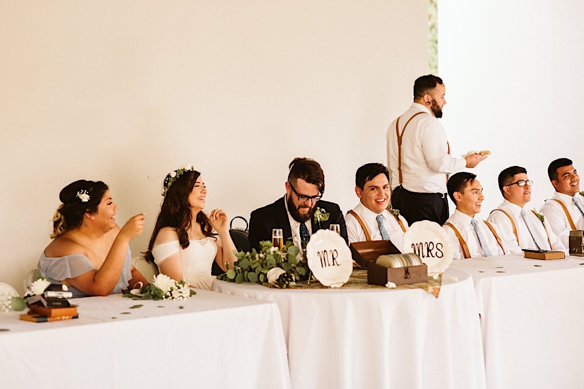 Bride and groom sit with wedding party at their reception behind "Mr" and "Mrs" platters. White and green bouquet on table.