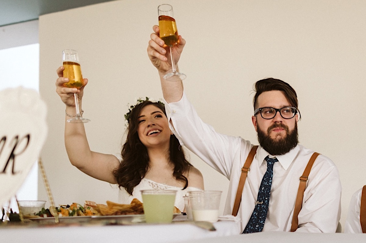 Bride and groom sit at a table and raise their champagne glasses in a toast