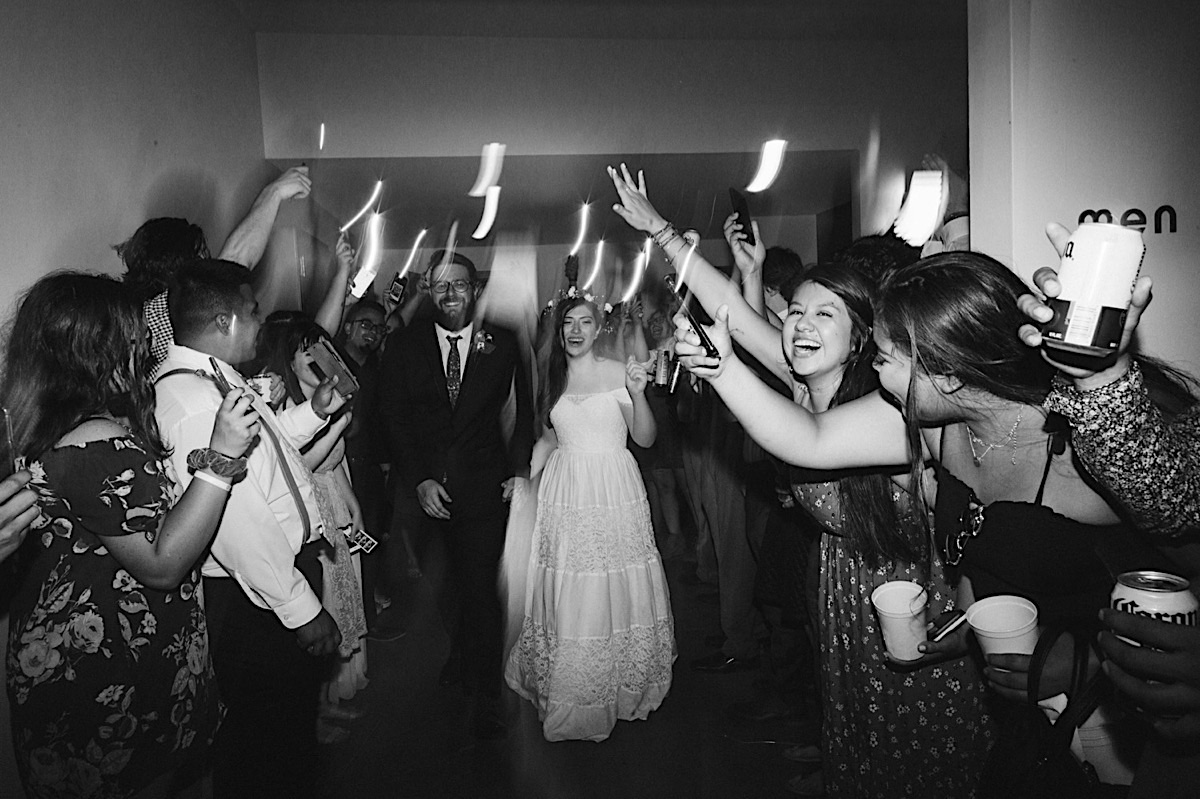 Bride and groom exit through a makeshift "sparkler" tunnel. Guests hold cellphones to light their way.
