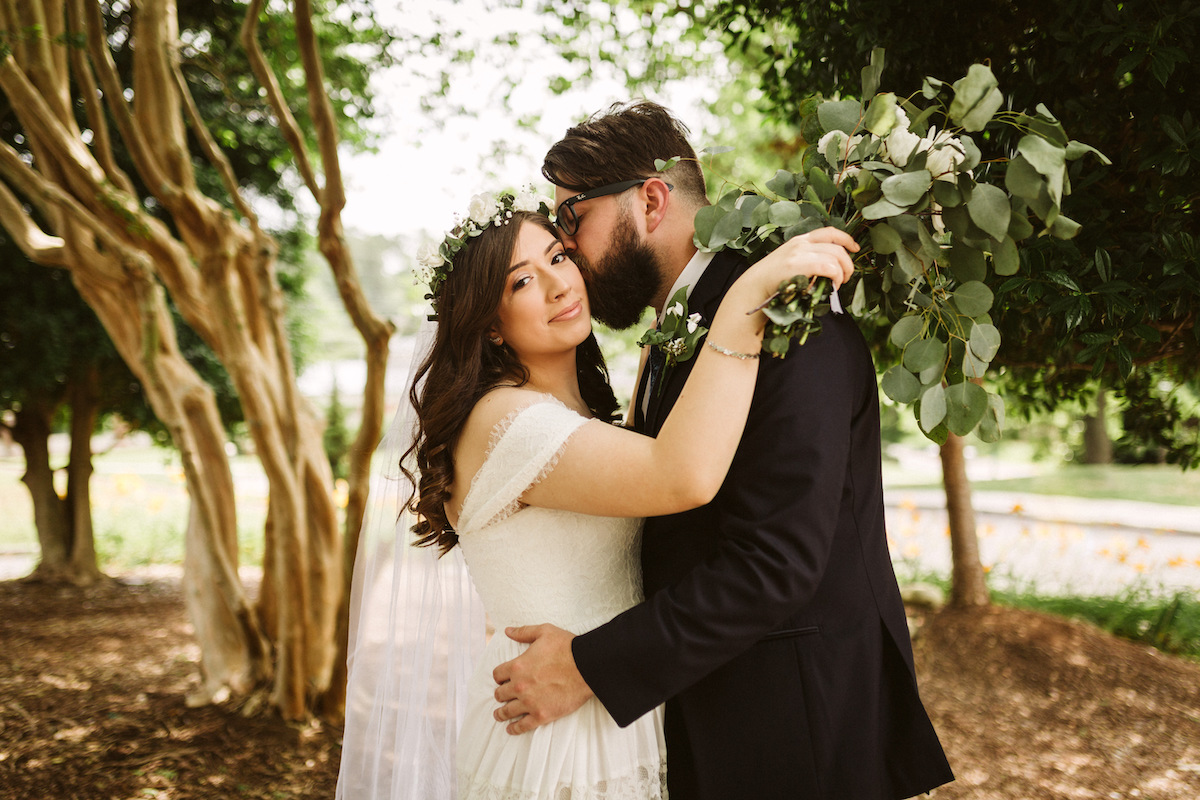 Bride looks at the camera while groom kisses her cheek. Her white floral bouquet cascades over his shoulder.