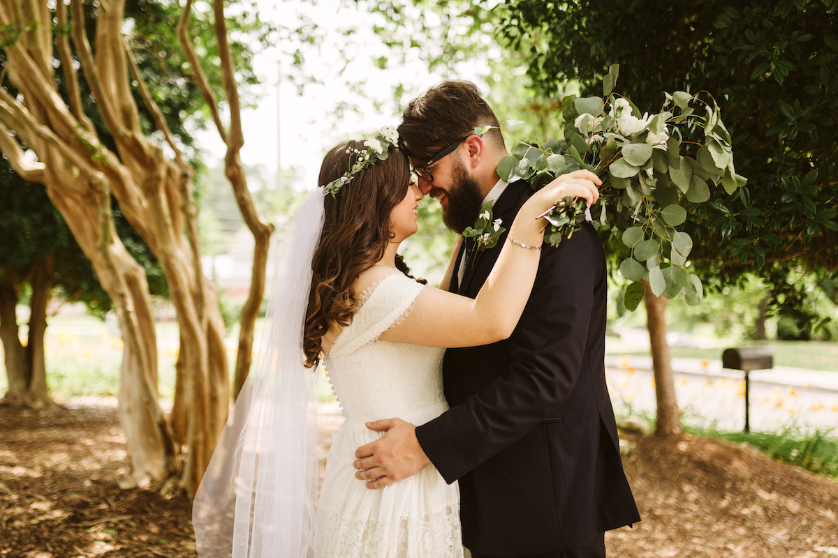 Bride and groom stand facing each other, cuddling their noses to each other. Her white bouquet falls over his shoulder.