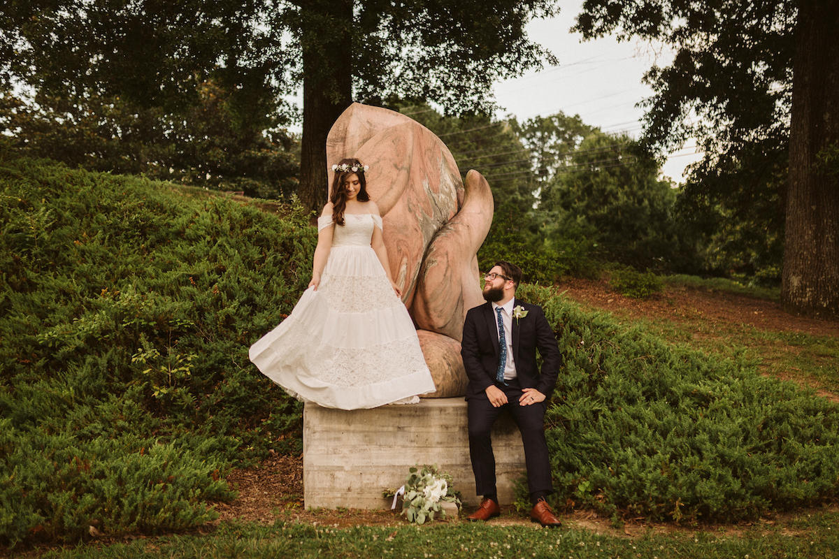 Groom sits looking up at bride who is standing on statue flaring her skirt at Creative Arts Guild wedding in Dalton, Georgia.