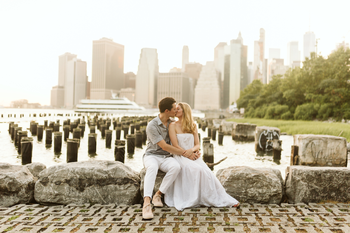 Man and woman sit on large rocks near water with New York City scape behind them