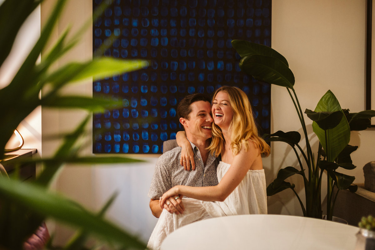 Woman sits on man's lap in front of a large black and blue painting as they laugh. They're surrounded by large plants.